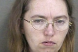 A woman with mousey hair, glasses and weak chin scowls at the camera in a police mug shot.