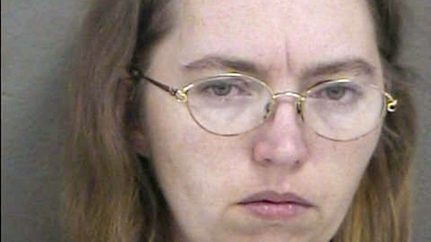 A woman with mousey hair, glasses and weak chin scowls at the camera in a police mug shot.