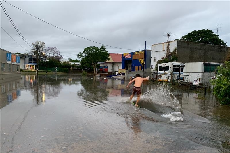 A shirtless man balances on a board, surfing through floodwater that appears to be a foot deep. It covers the whole road
