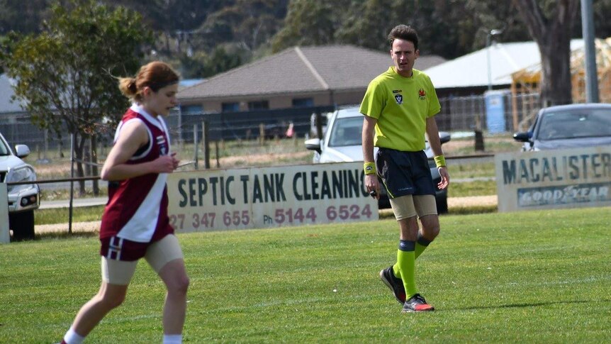 An umpire looks on as a female footballer jogs past.