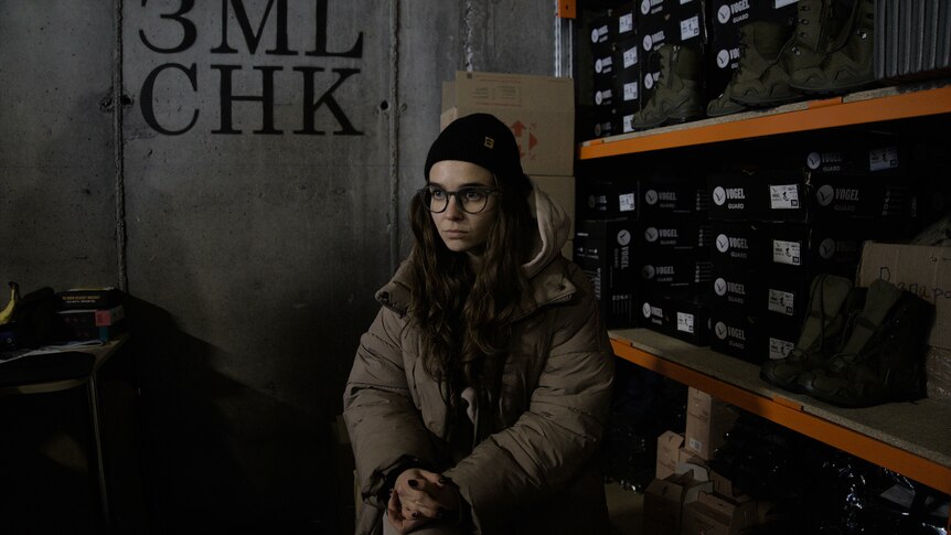 A woman with long hair and glasses stands next to a shelf of boots. 