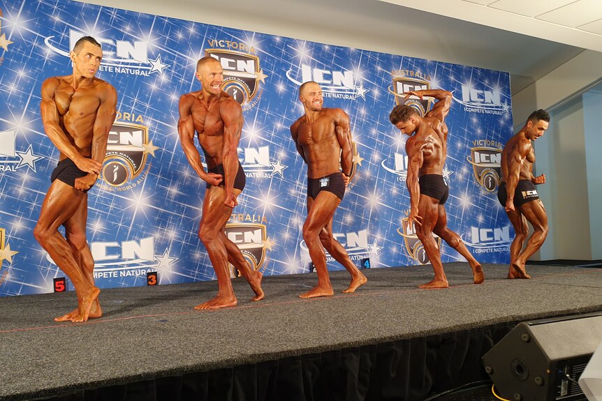 Five tanned, muscly bodybuilder men, including Matthew Nunan pose on stage