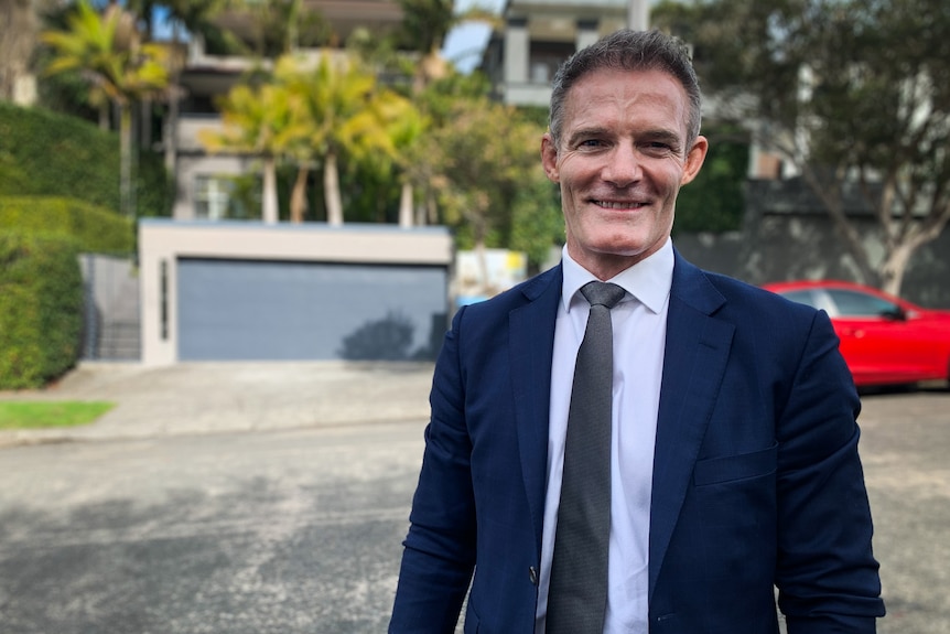 A man wearing a dark blue suit and tie, standing in front of a home for sale, smiles at the camera