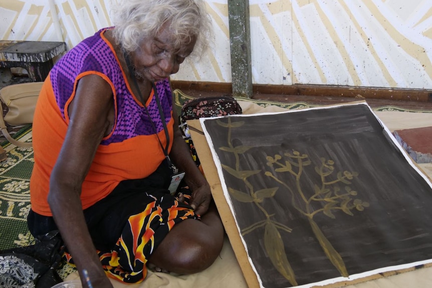 An Aboriginal woman with white hair sits on a rug, painting a piece of canvas.