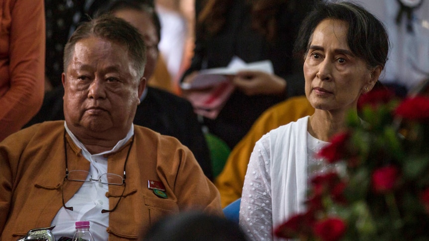 A man in saffron jacket sitting next to Aung San Suu Kyi in white.
