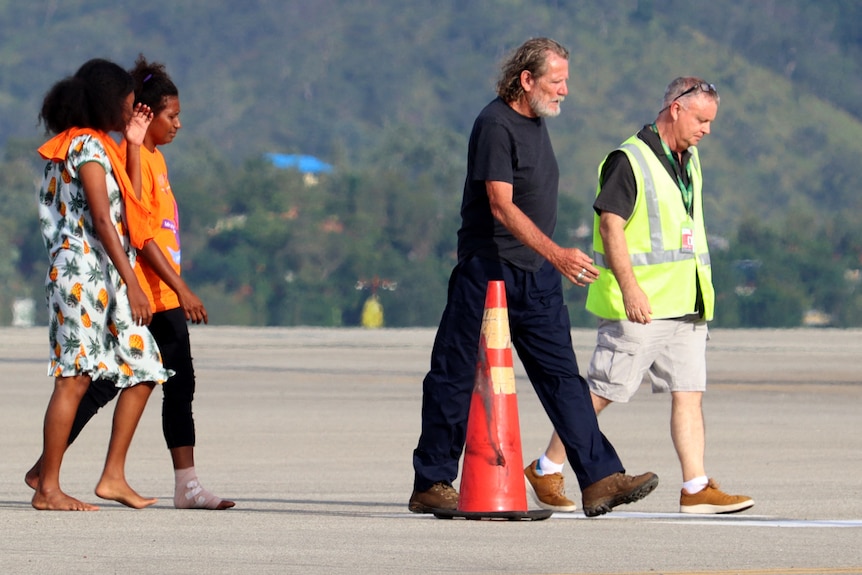 A man wearing black jeans and dark shirt walks on the tarmac in front of two women