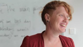 A woman smiling broadly stands before a blurred whiteboard with hands outstretched, looking to side.