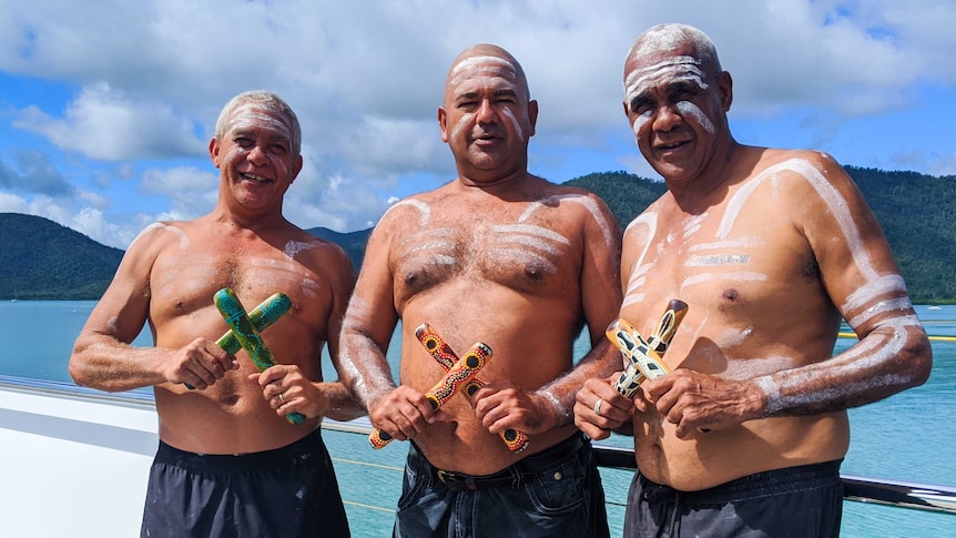 Three men painted with traditional ochre holding clap sticks standing in front of Whitsundays islands and blue water