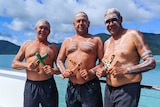 Three men painted with traditional ochre holding clap sticks standing in front of Whitsundays islands and blue water