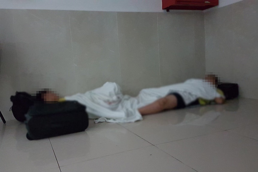 Two young boys sleeping on a floor with white blankets over them and using backpacks as pillows