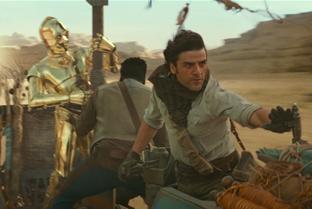 A gold droid and man cling on as a man wearing dark gloves and scarf pilots a desert craft at high speed.