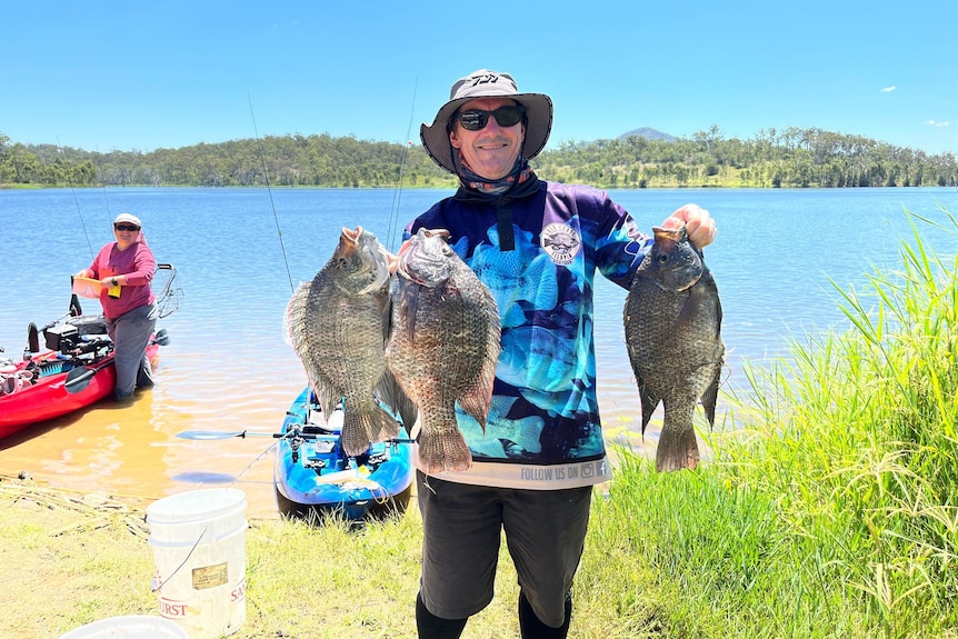 A fisherman holds up three large fish with a lake and another fisher behind him.