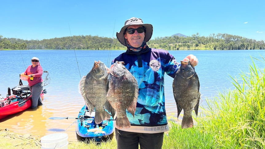 A fisherman holds up three large fish with a lake and another fisher behind him.