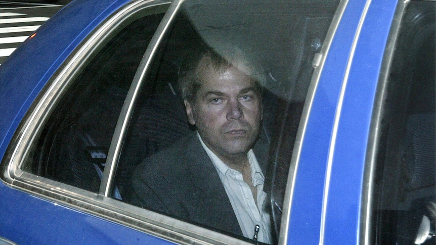 A man wearing a suit is seen looking solemn through the window of a car