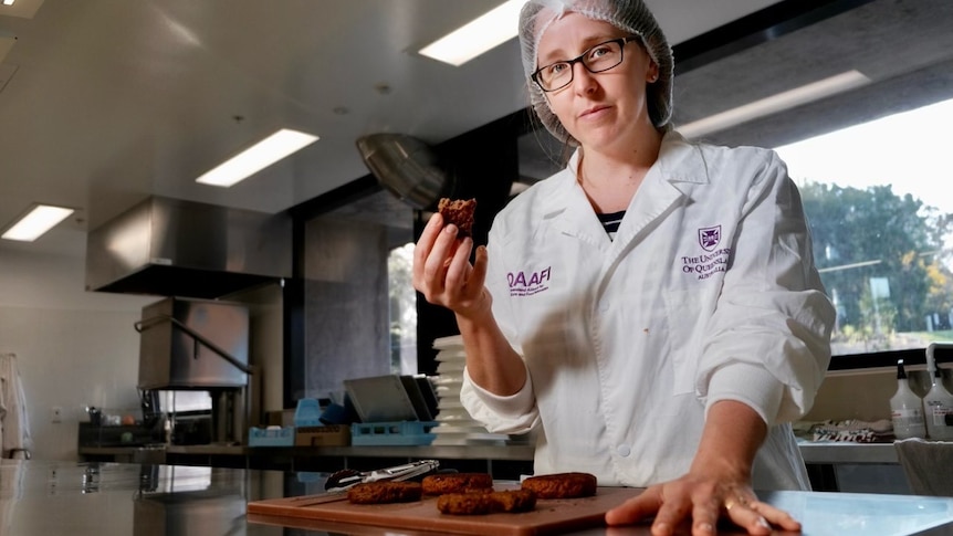 Heather Smyth in a white lab coat and hair net, hand on bench, fake meat on chopping board, holding fake meat.