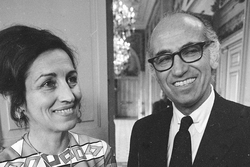 A woman in a sixties style dress with dark hair stands next to a man in a suit and glasses smiling at camera