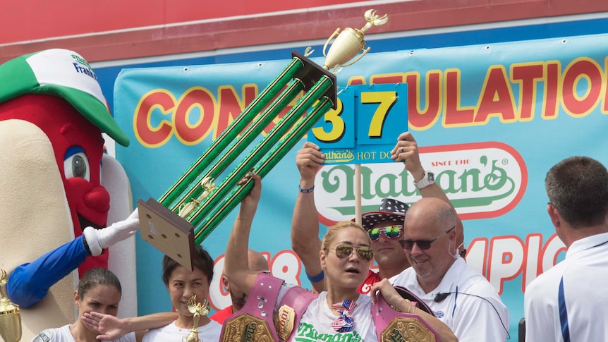 Miki Sudo holds her trophy up and is draped in her four previous championship belts