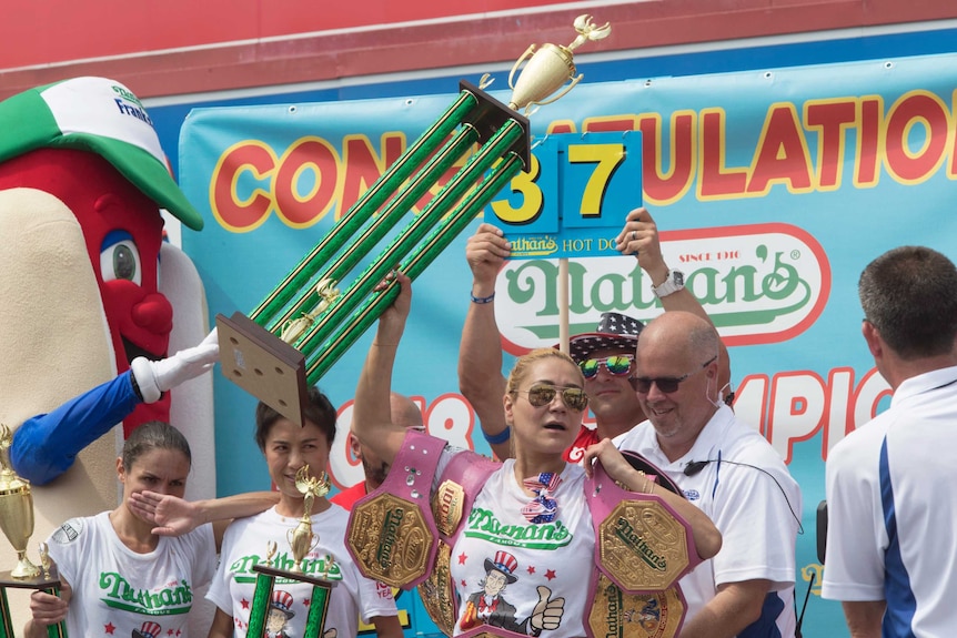 Miki Sudo holds her trophy up and is draped in her four previous championship belts