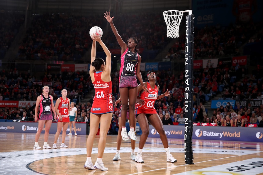 A Super Netball defender leaps high with arm outstretched to block a shot from an opposition player near the basket.