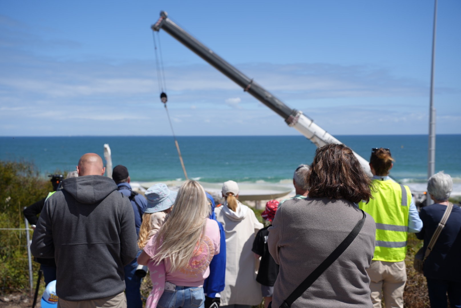 A crowd of people stands looking towards a crane on City Beach on a sunny day.