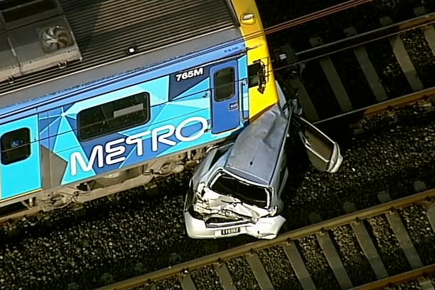 A silver car crumpled underneath a blue and yellow Metro train on the train tracks.