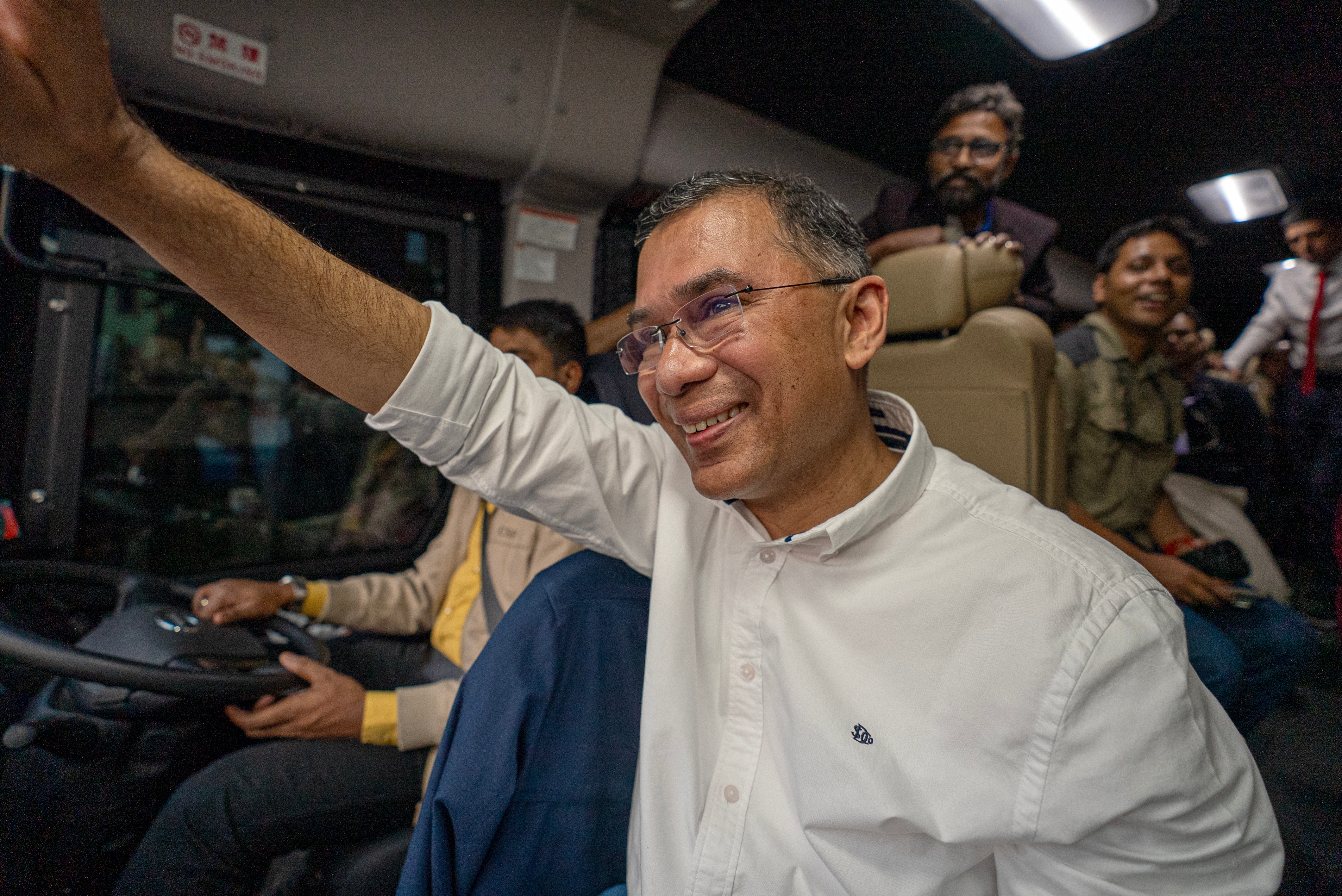 A Bangladesh man raises his arm, smiling and waving in a bus with other supporters.
