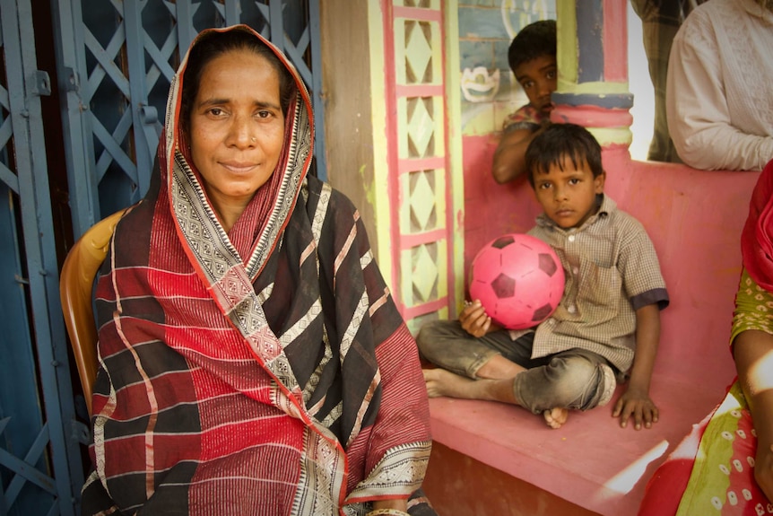 Anwoara sits in front of a blue door with two children in the background, one holding a pink soccer ball.