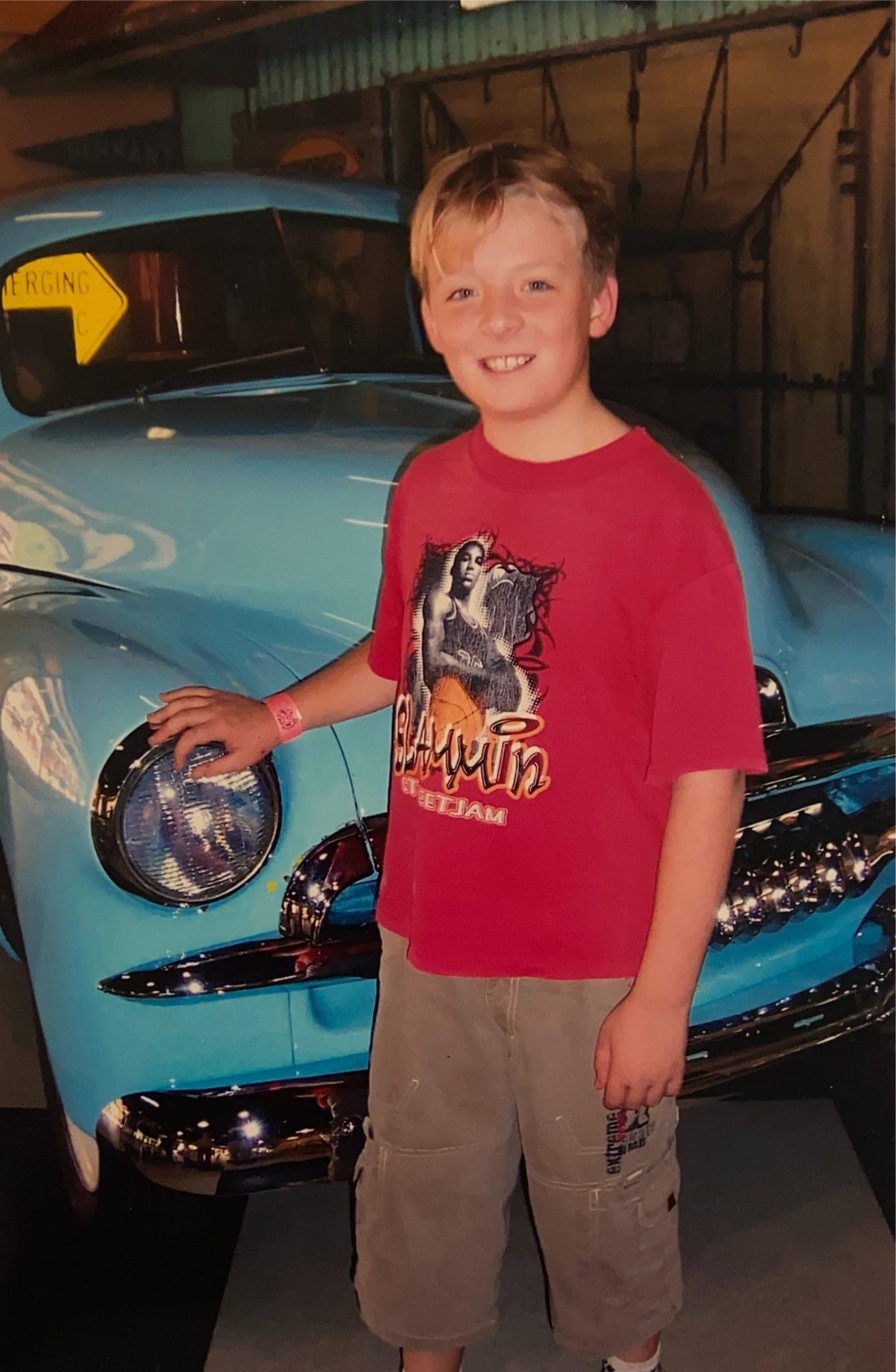 A boy in a red t-shirt stands next to a pale blue vintage car