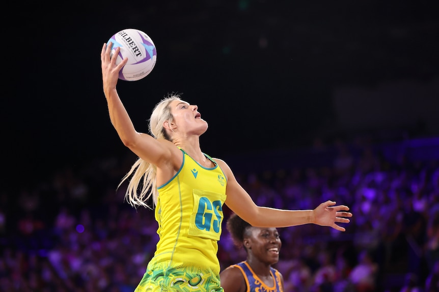A netball player wearing yellow and green jumps with the ball