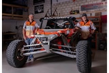 two men stand either side of a bright orange, off-road buggy. They are wearing matching bright orange shirts and black caps