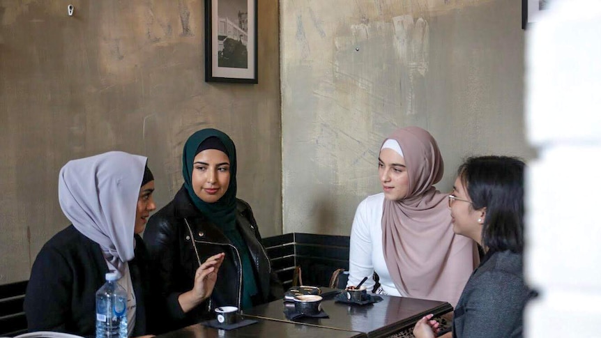 Four women sit in a cafe talking, three are wearing headscarves and one is not.