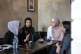 Four women sit in a cafe talking, three are wearing headscarves and one is not.