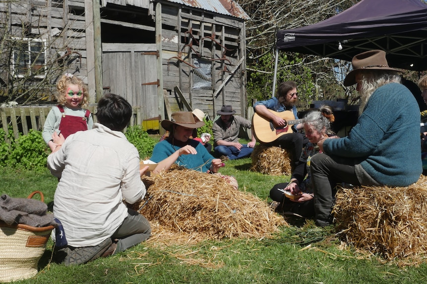 A family sits on hay bales in the sun eating lunch while a busker plays a guitar in the background 