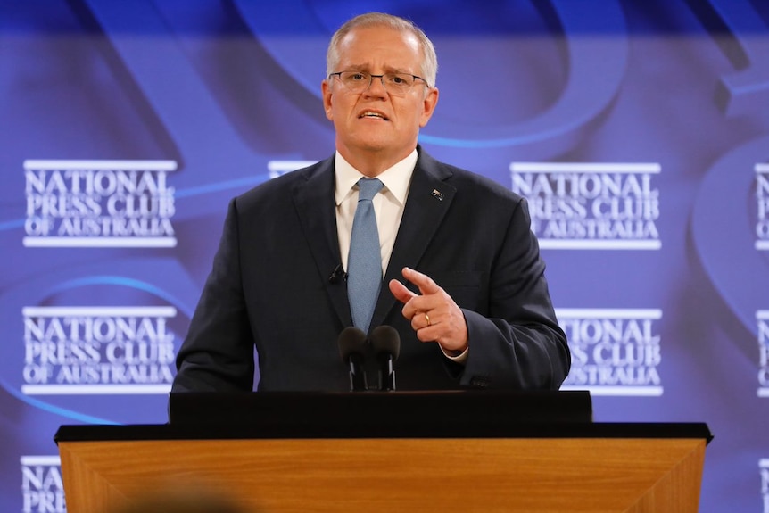 Un homme à lunettes se tient devant un lutrin devant un fond bleu pendant qu'il prononce un discours.