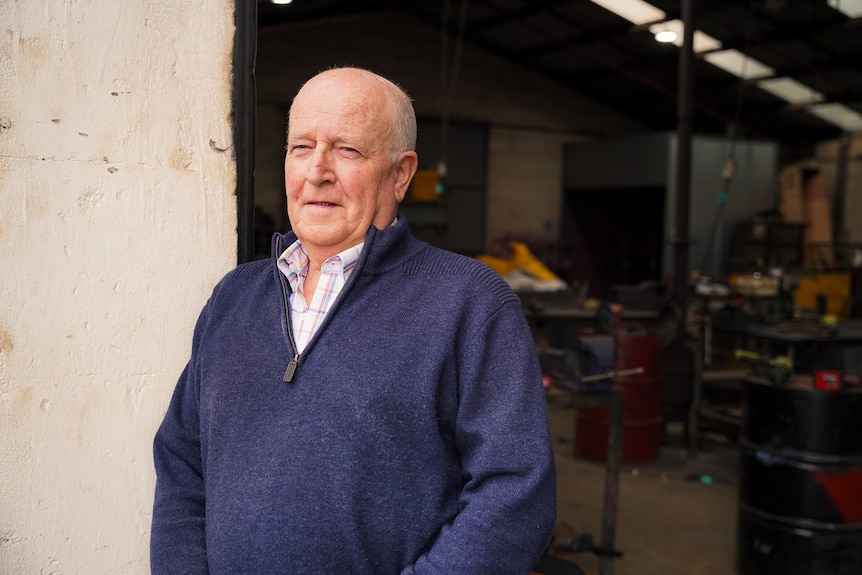 A man with a bald head and navy sweater stands in a large doorway of a workshop.