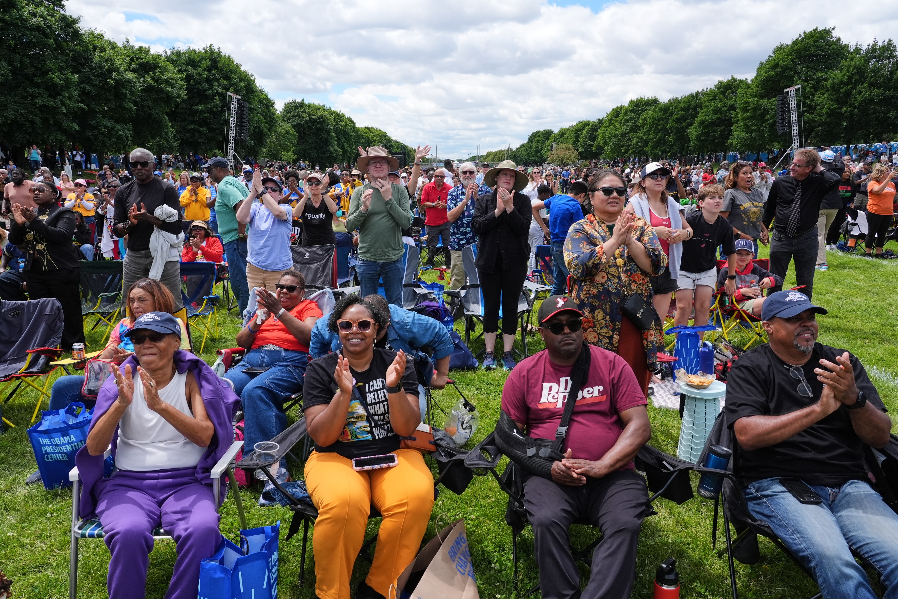 A crowd of people stand and sit on camp chairs in a park.