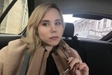 A young blonde woman in a camel coat and black nail polish takes a selfie in a car