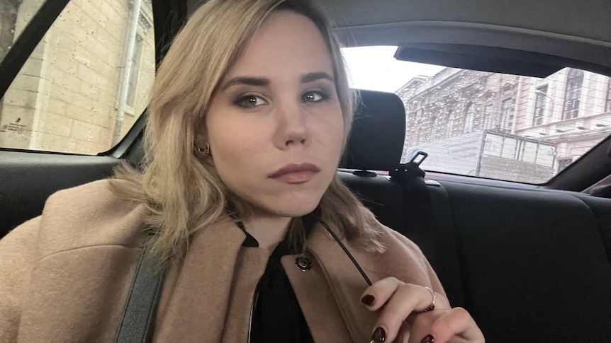 A young blonde woman in a camel coat and black nail polish takes a selfie in a car