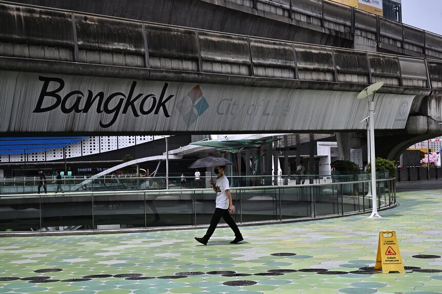 A pedestrian walks under an umbrella at an overpass on which "Bangkok" is written, with no one else in the photo.