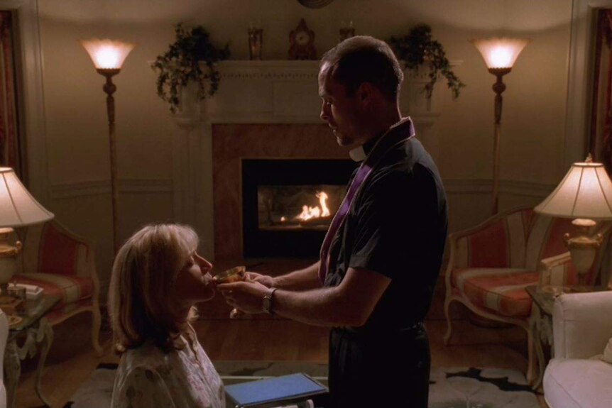 Sopranos priest Father Intintola giving Carmela Soprano wine in a chalice in front of a fireplace.