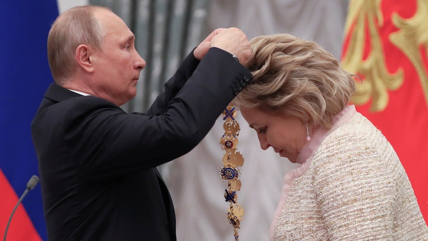 Vladimir Putin places a gold necklace around the neck of a blonde woman bowing her head