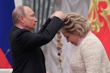 Vladimir Putin places a gold necklace around the neck of a blonde woman bowing her head
