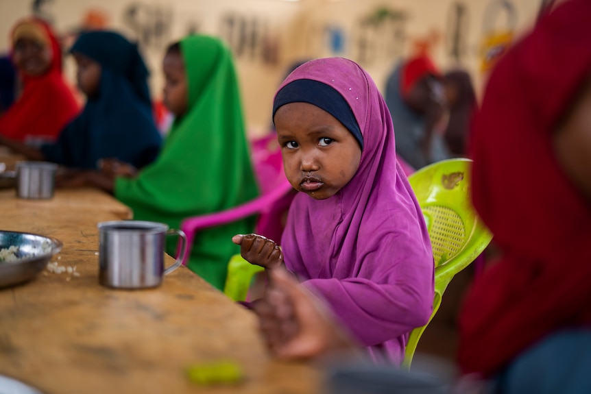a child in purple clothes holds food in their hand while sitting a school with other children