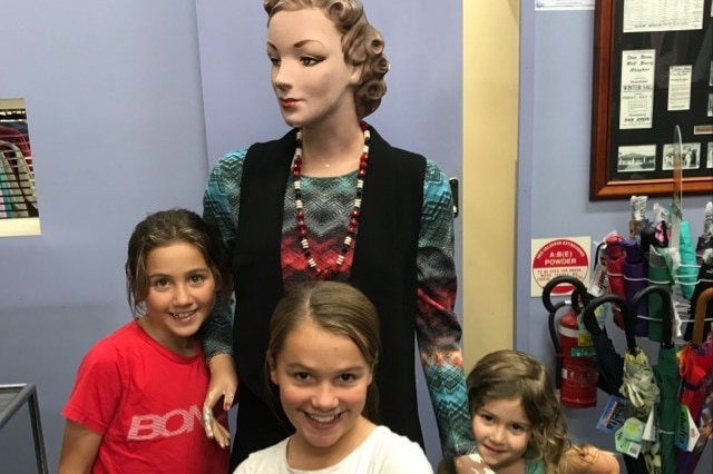 Three young girls stand next to a store mannequin in a clothing shop.