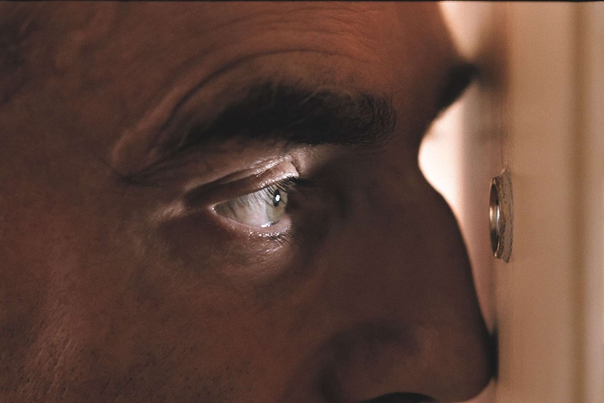 Daniel Day-Lewis, as Reynolds Woodcock, stares through the peep hole of a door in Phantom Thread.