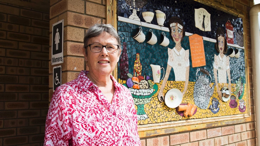 A woman stands with a mosaic of a cafe scene behind her.