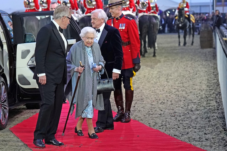 The Queen arrives, leaving a car with a walking stick on a red carpet. 