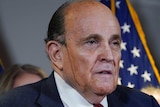 A man in a suit with sweat on his face has a line of black dripping down the side of his cheek as he stands at a podium