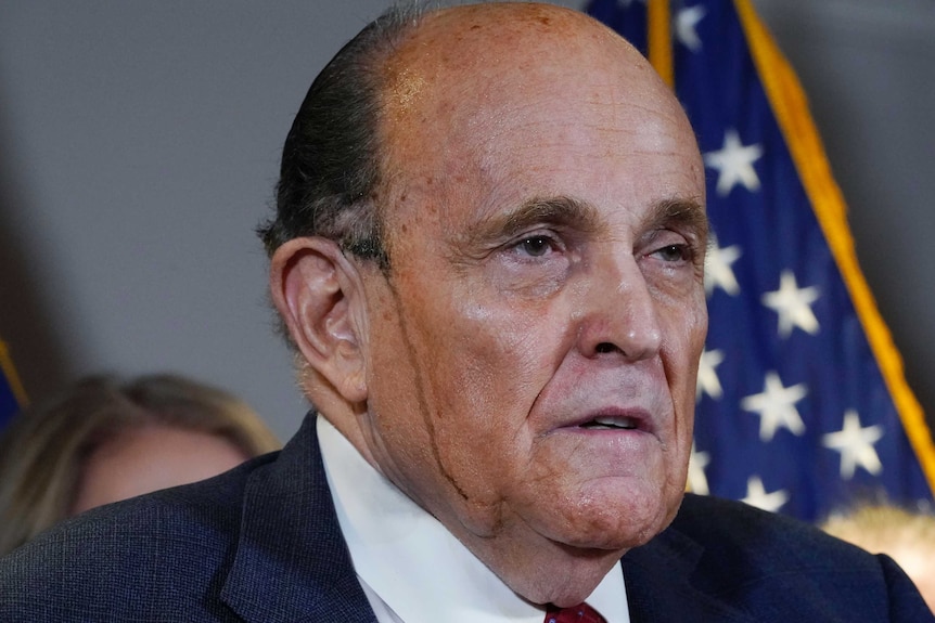 A man in a suit with sweat on his face has a line of black dripping down the side of his cheek as he stands at a podium