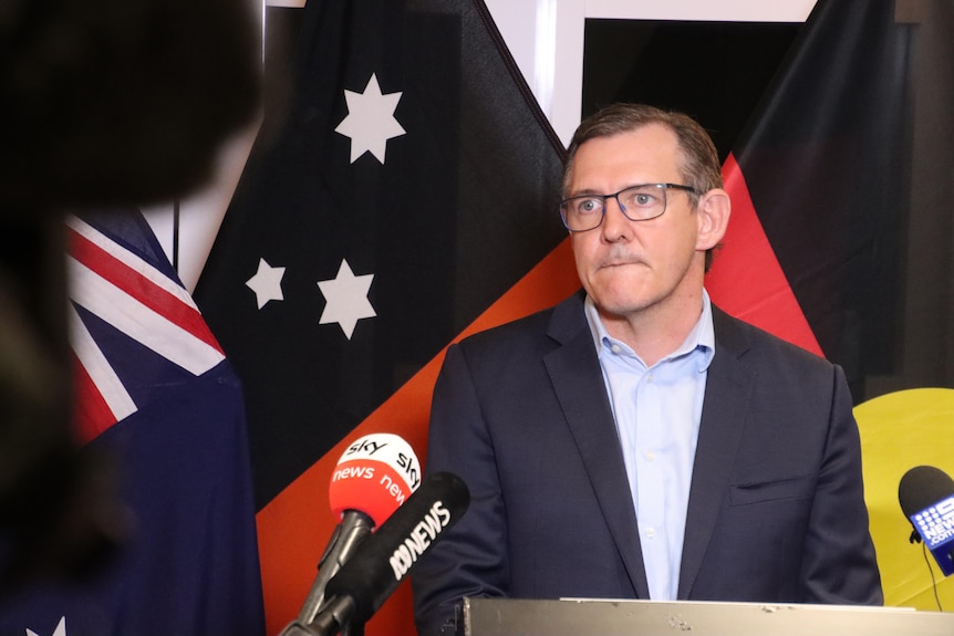 Un homme aux cheveux noirs et à lunettes dans un costume sombre – le ministre en chef du NT, Michael Gunner – parle devant un drapeau australien. 